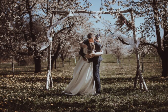 Eine Braut und ein Bräutigam küssen sich unter einer Laube in einem Obstgarten.