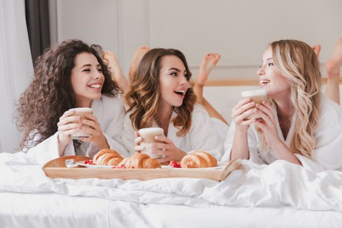 Drei Frauen in Roben sitzen auf einem Bett mit Kaffee und Croissants.