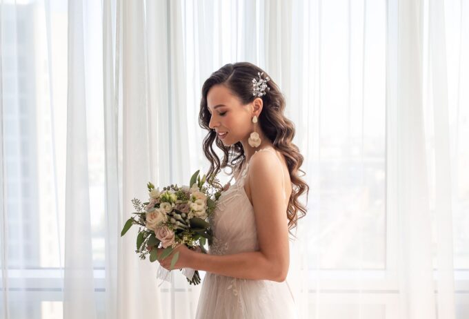 Eine Braut hält einen Blumenstrauß vor einem Fenster.