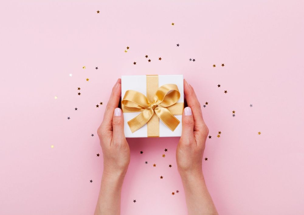 Hände halten eine Geschenkbox mit goldenem Band auf rosa Hintergrund.