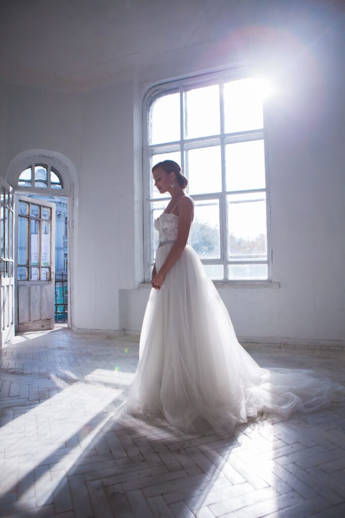 Eine Braut in einem Hochzeitskleid steht in einem Raum mit Sonnenlicht.