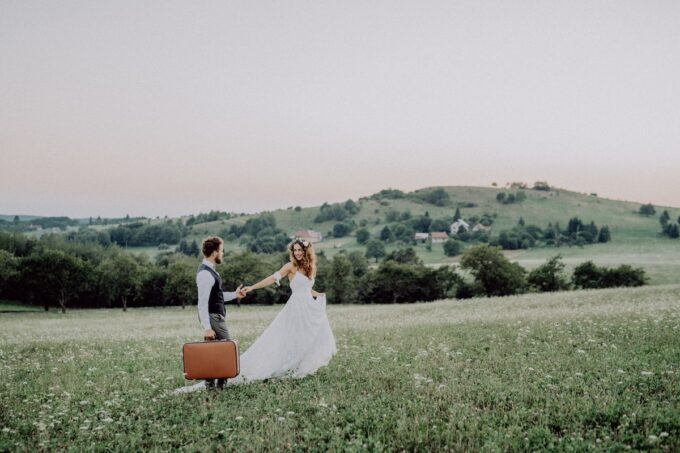 Eine Braut und ein Bräutigam halten bei Sonnenuntergang Koffer auf einem Feld.