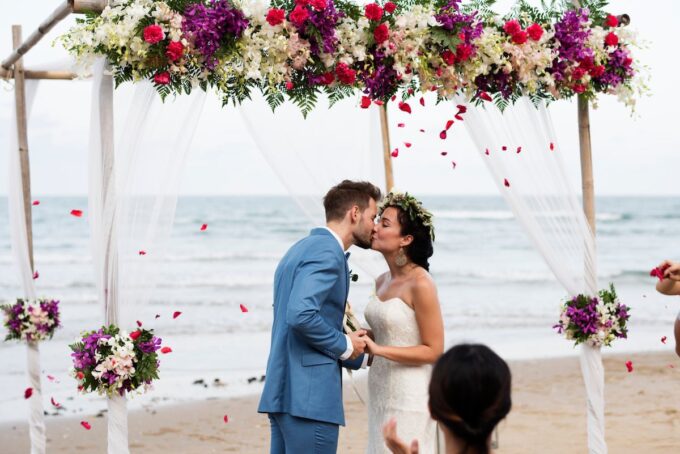 Eine Braut und ein Bräutigam küssen sich unter einem Bogen am Strand.