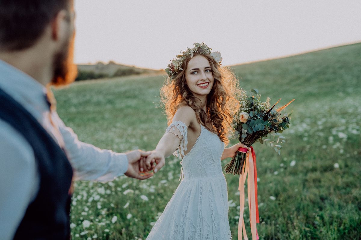 Eine Braut und ein Bräutigam halten sich bei Sonnenuntergang auf einem Feld an den Händen.