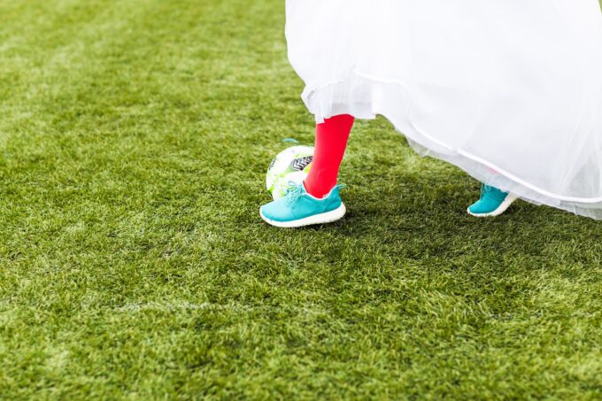 Eine Frau in einem Hochzeitskleid läuft mit einem Fußball über Gras.
