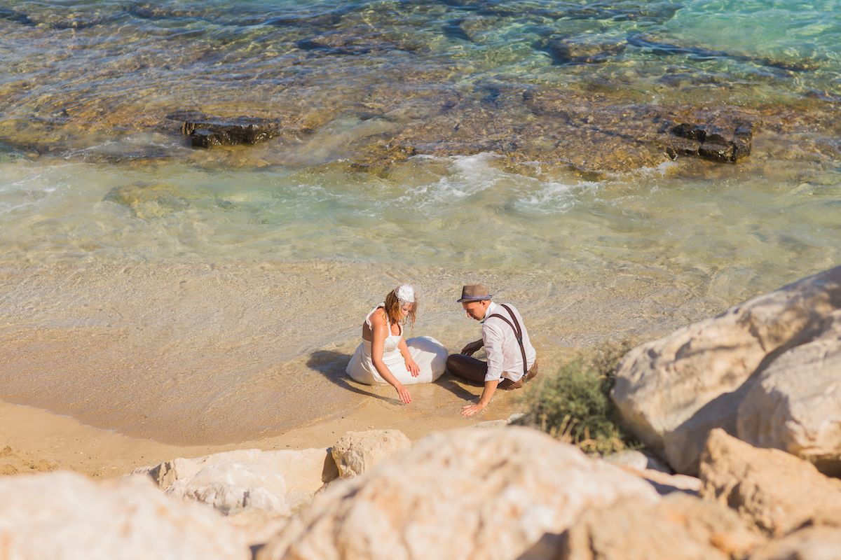 Eine Braut und ein Bräutigam kniend im Sand neben dem Wasser.