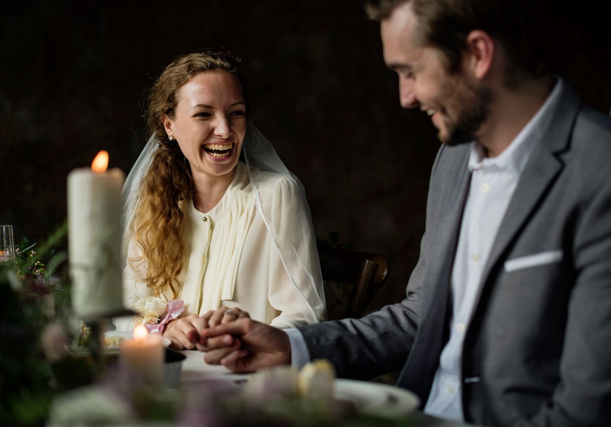 Eine Braut und ein Bräutigam lachen an einem Esstisch.
