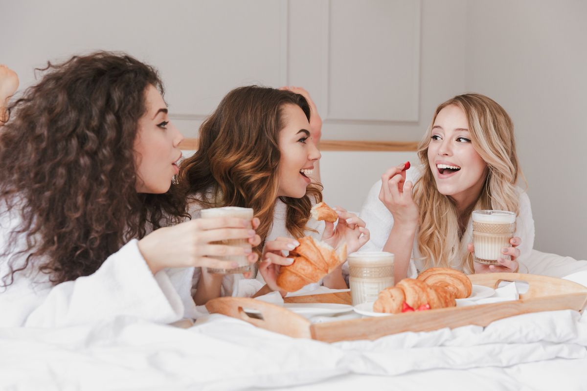 Drei Frauen in Roben trinken Kaffee und essen Croissants im Bett.