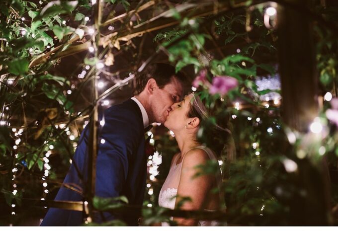 Eine Braut und ein Bräutigam küssen sich unter Lichtern in einem Garten.