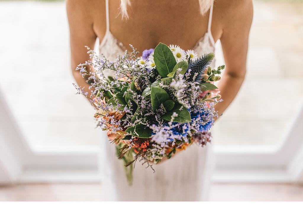 Eine Braut hält einen Strauß Wildblumen vor einem Fenster.