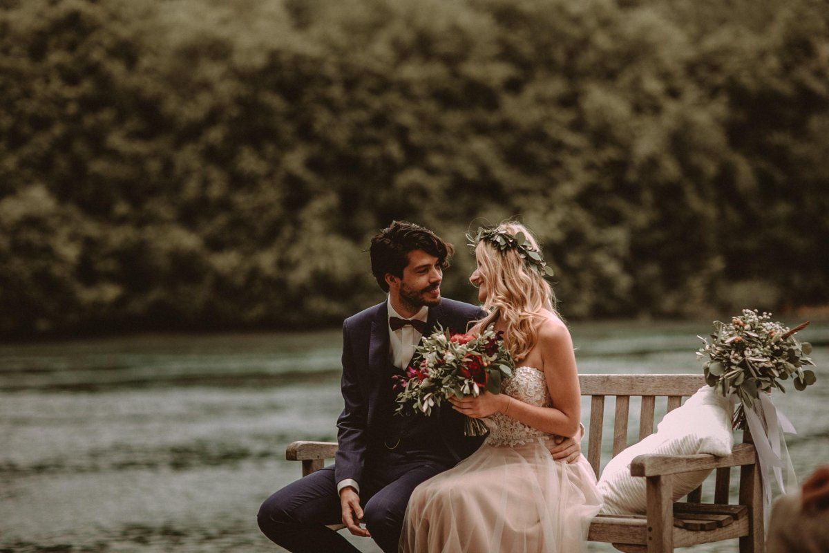 Eine Braut und ein Bräutigam sitzen auf einer Bank neben einem Fluss.