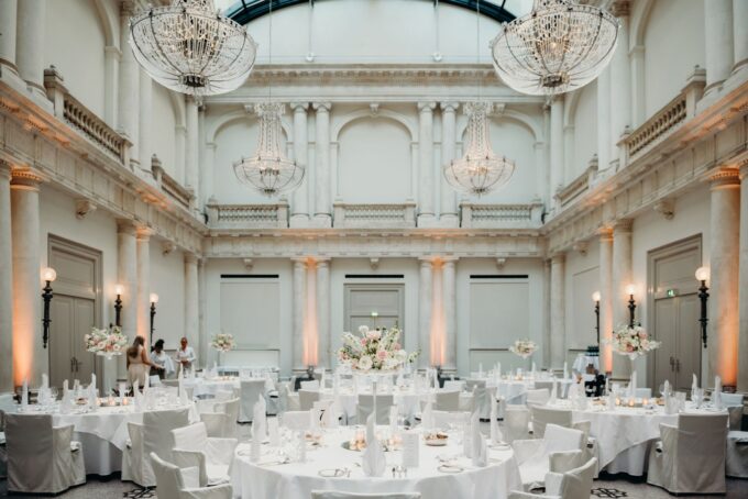 Eine Hochzeitsfeier in einem großen Saal mit weißen Tischen und Kronleuchtern.