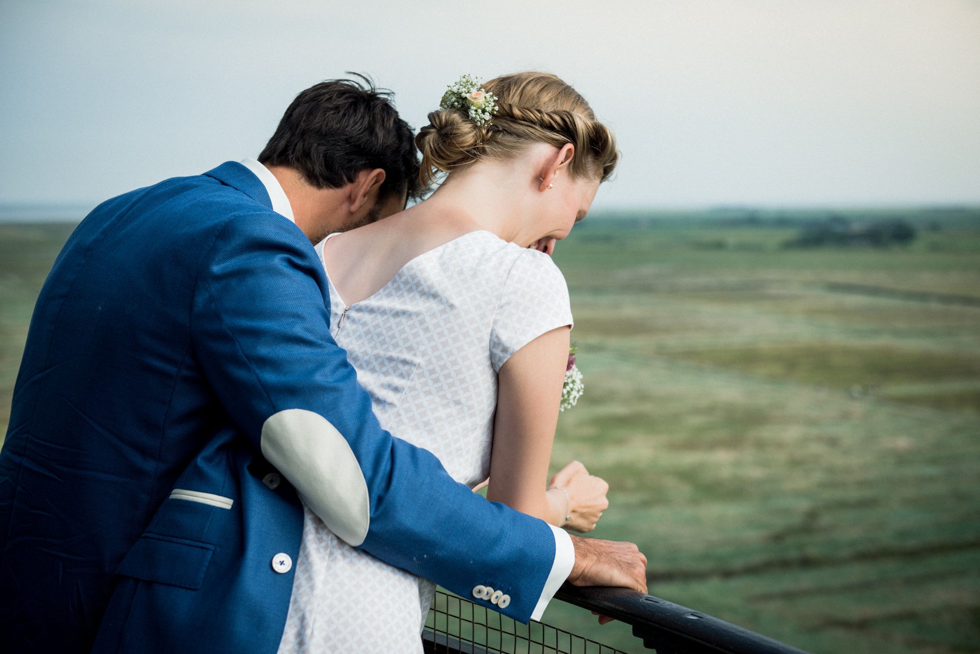 Eine Braut und ein Bräutigam umarmen sich auf einem Geländer mit Blick auf ein Feld.