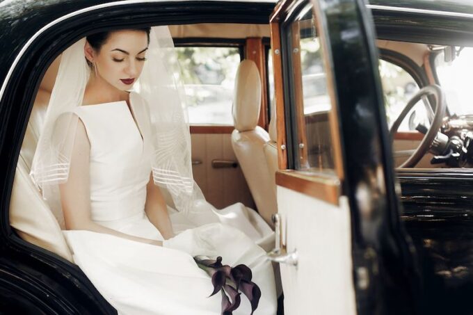 Eine Braut im Hochzeitskleid sitzt auf dem Rücksitz eines Oldtimers.