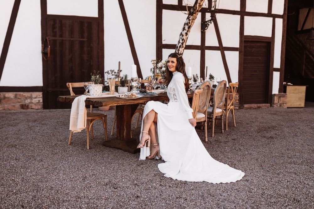Eine Braut in einem weißen Kleid sitzt an einem Tisch vor einer Scheune.