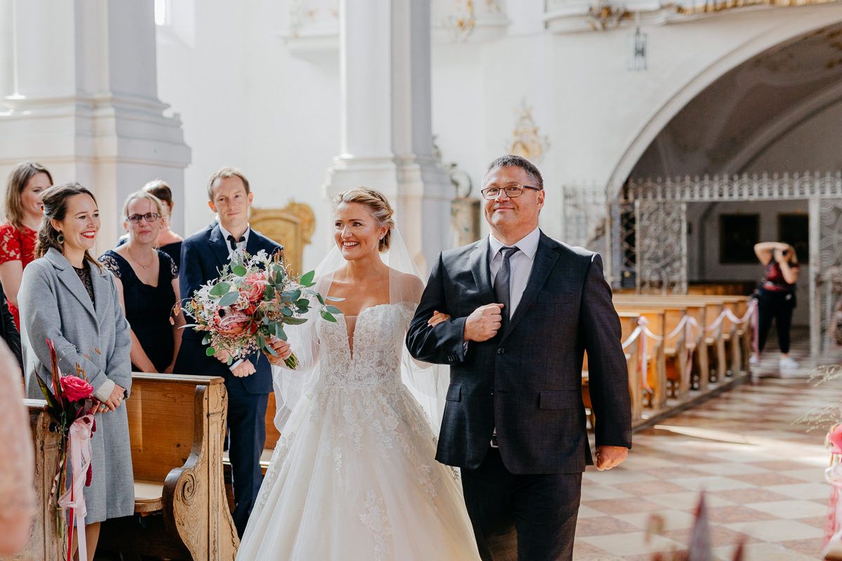 Eine Braut und ihr Vater gehen in einer Kirche den Gang entlang.