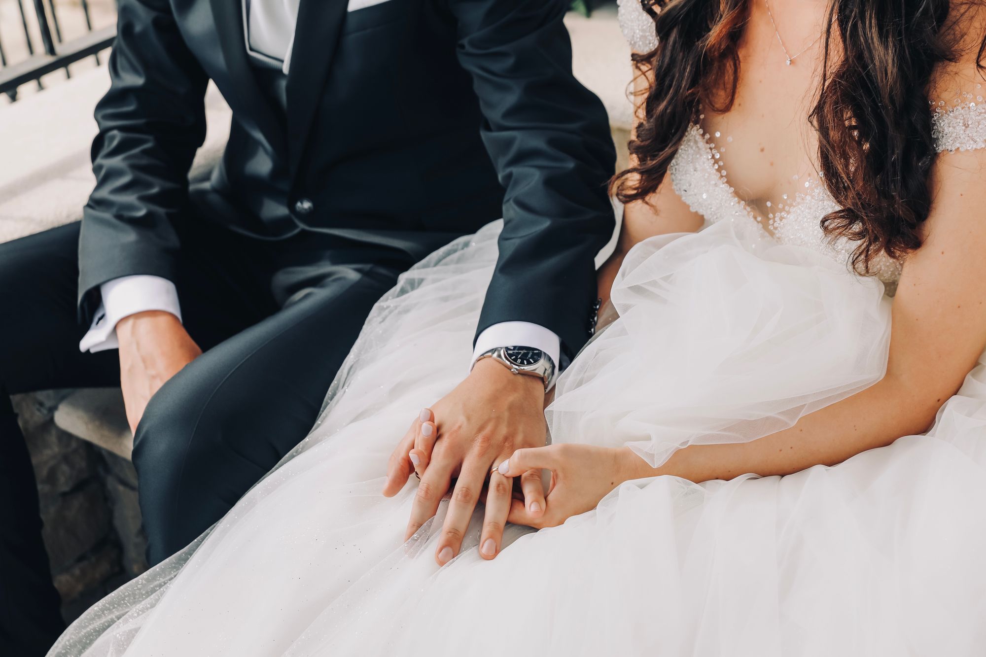 Eine Braut und ein Bräutigam sitzen Händchen haltend auf einer Bank.