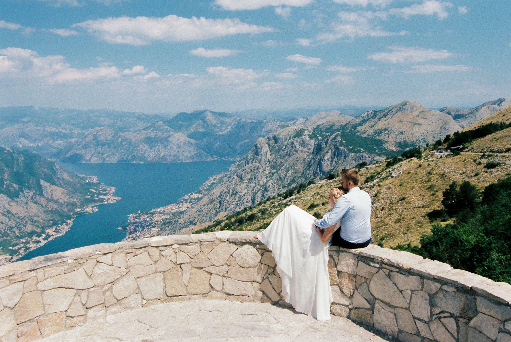 Eine Braut und ein Bräutigam sitzen auf einer Steinmauer mit Blick auf einen See.