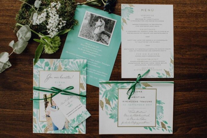 Hochzeitseinladungen mit grünen Blättern und Moos auf einem Holztisch.