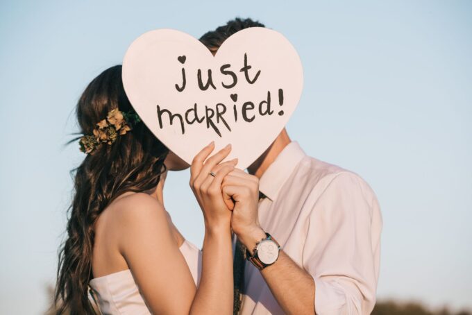 Eine Braut und ein Bräutigam küssen sich vor einem herzförmigen Schild mit der Aufschrift „Frisch verheiratet“.