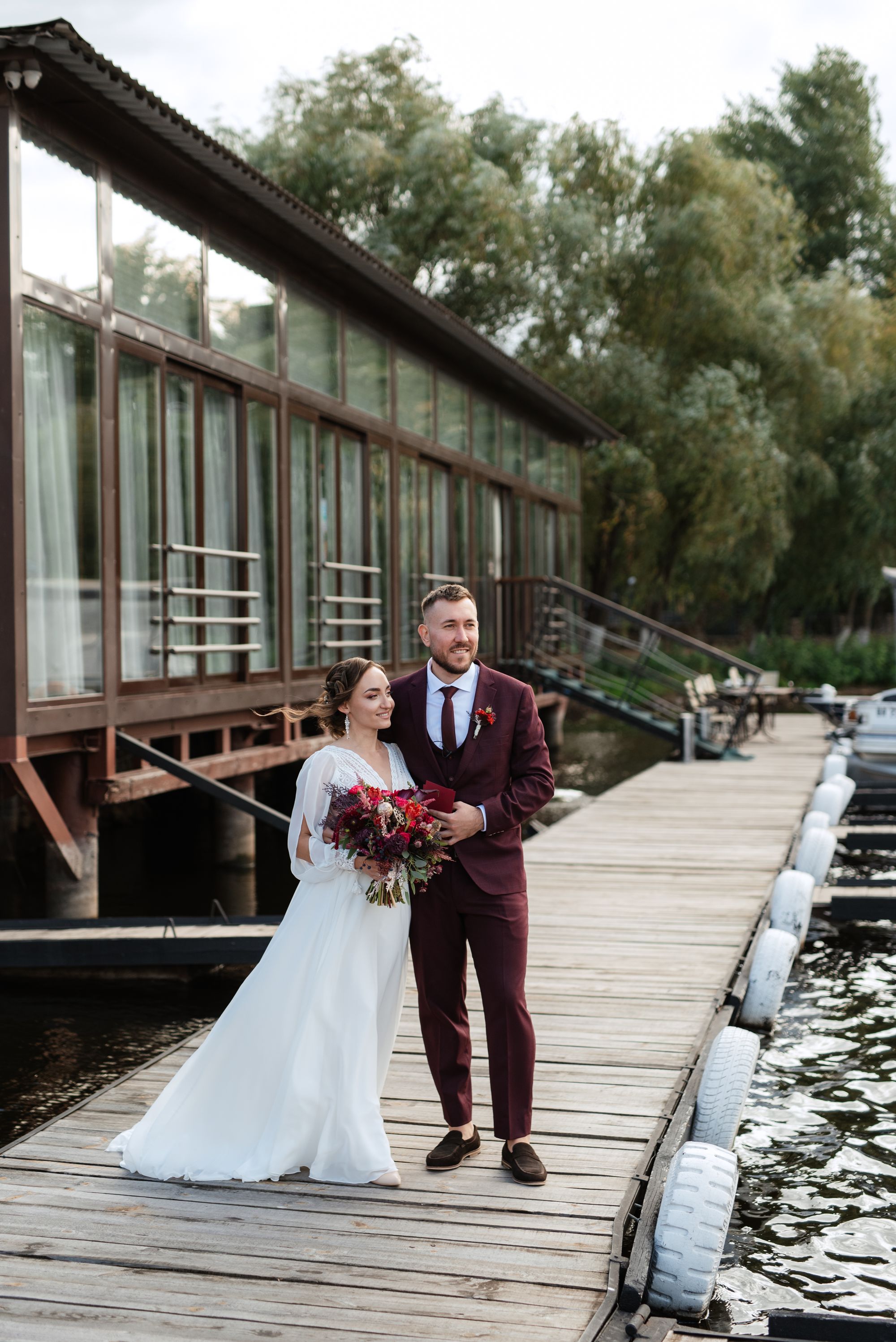 Brautpaar steht auf einem Steg - der Bräutigam trägt einen weinroten Anzug