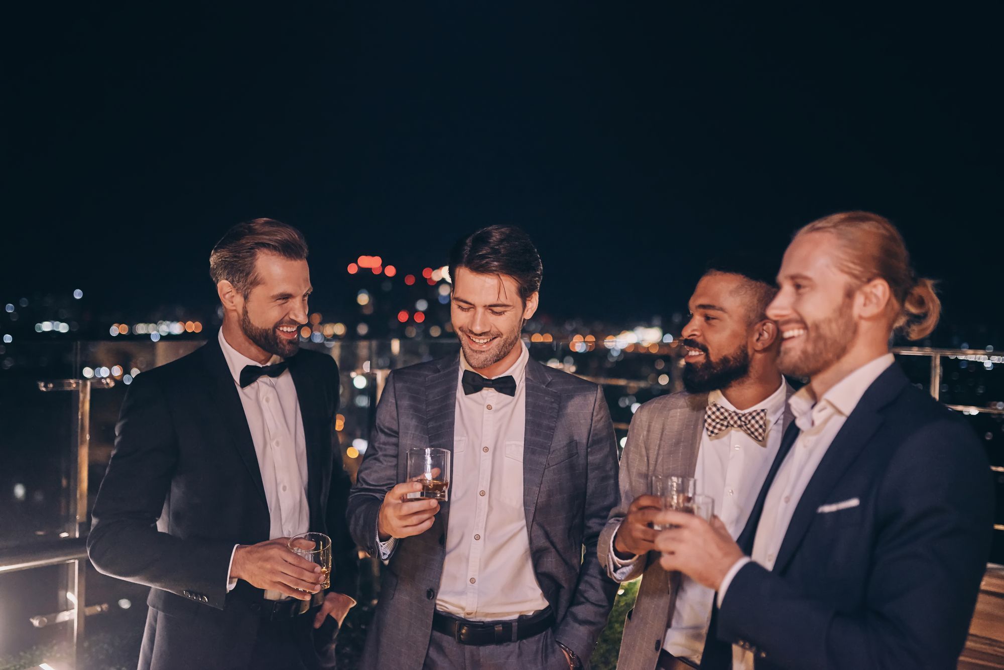 Vier Männer im Smoking trinken nachts Wein auf einem Dach.