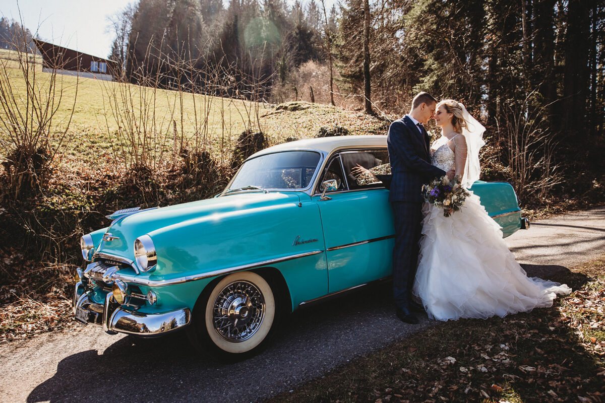 Eine Braut und ein Bräutigam posieren neben einem blauen Oldtimer.