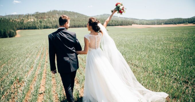 Brautpaar in Hochzeitsoutfits läuft eingehakt ein Feld entlang