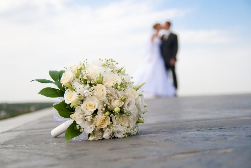 Im Vordergrund liegt der Brautstrauß auf dem Boden. Im Hintergrund küsst sich das Brautpaar.