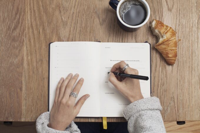 Die Hand einer Frau schreibt in ein Notizbuch mit einer Tasse Kaffee.