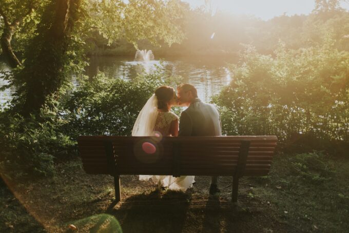 Eine Braut und ein Bräutigam küssen sich auf einer Bank vor einem See.