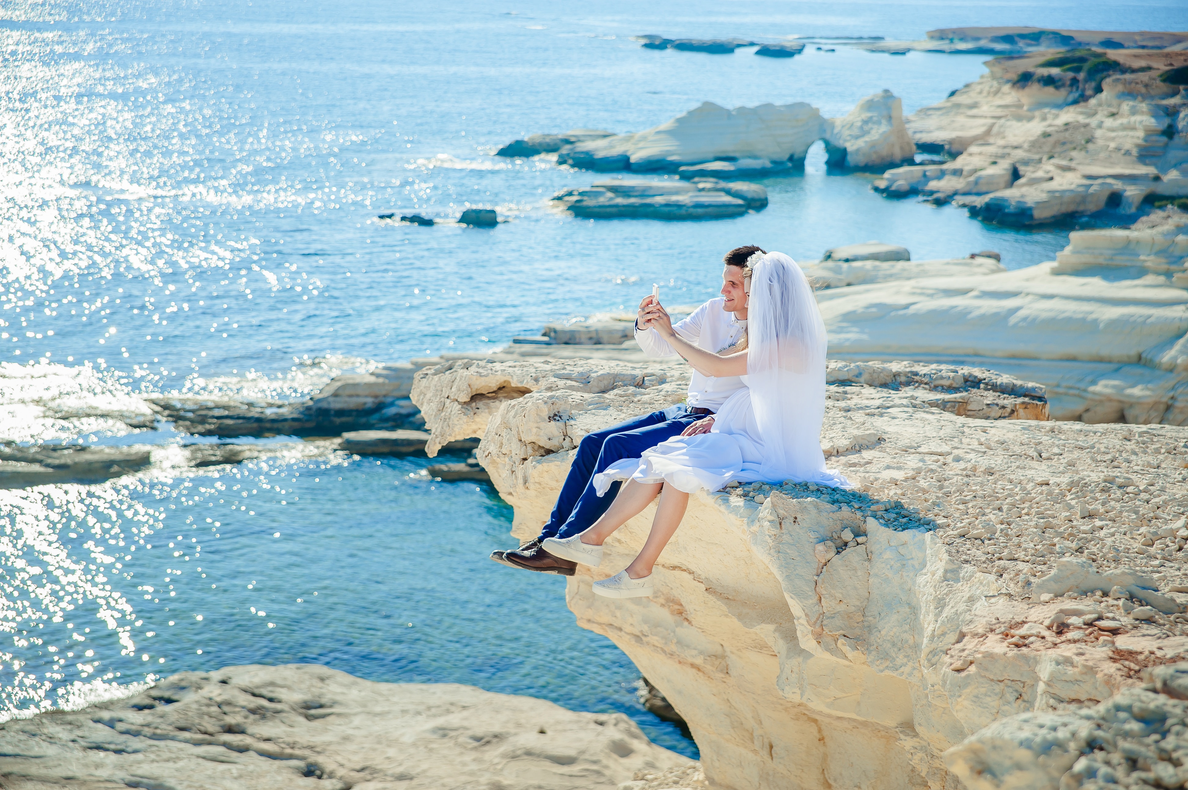 Eine Braut und ein Bräutigam sitzen auf einer Klippe mit Blick auf das Meer.