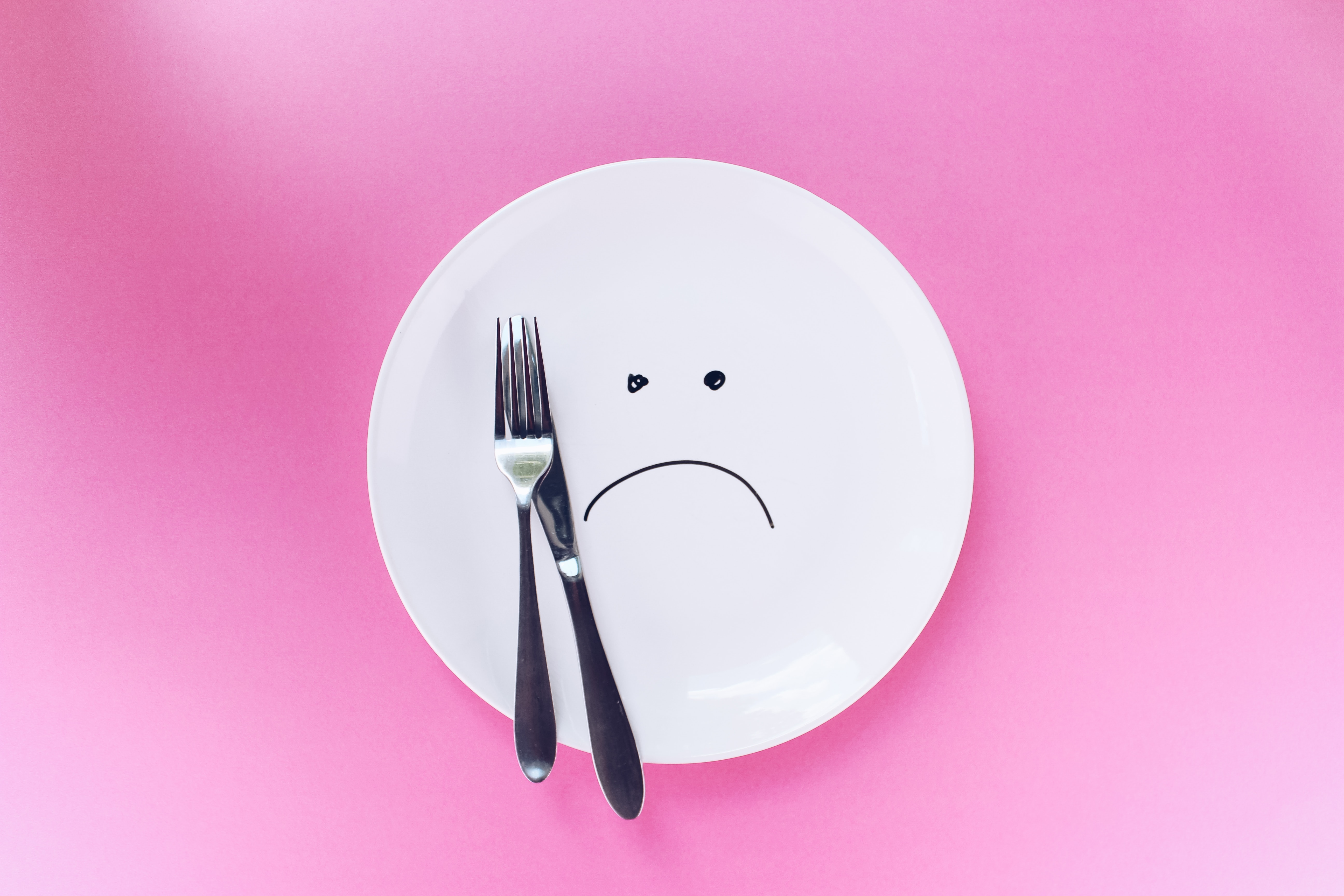 Ein Teller mit traurigem Gesicht und Gabel auf rosa Hintergrund.