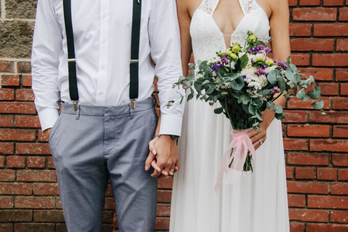 Eine Braut und ein Bräutigam halten sich an den Händen vor einer Ziegelwand.