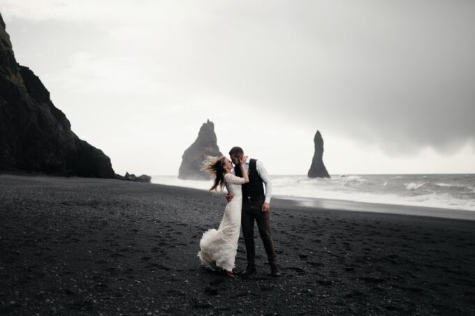 Eine Braut und ein Bräutigam küssen sich an einem schwarzen Sandstrand in Island.