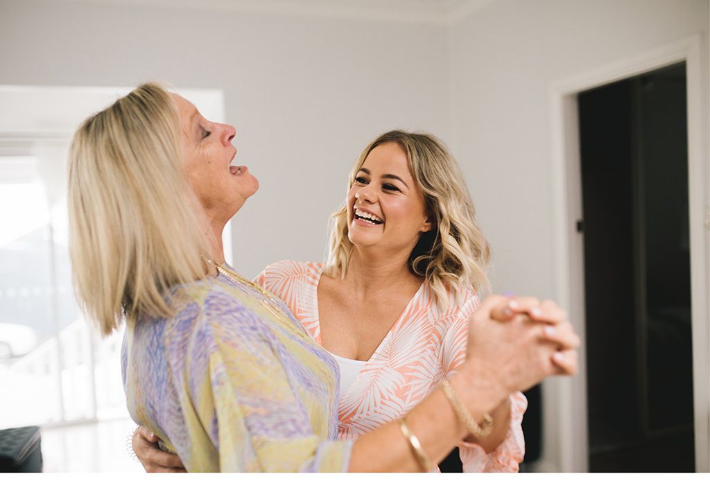 Zwei Frauen lachen und tanzen in einem Wohnzimmer.